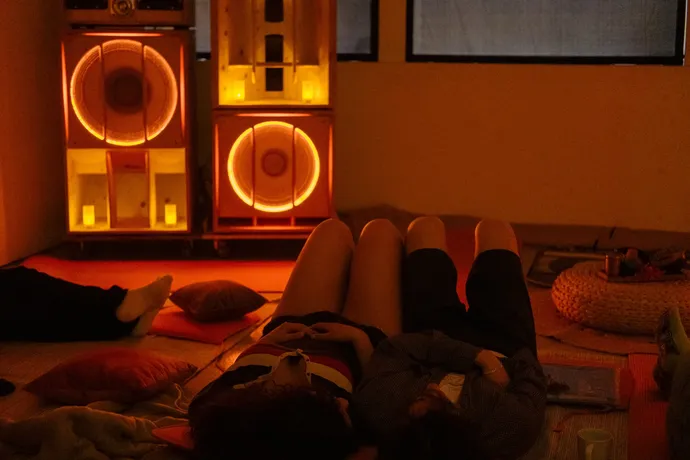 People relaxing on the floor with dim, warm lighting and glowing speakers in the background, creating a cozy atmosphere.