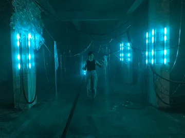 A person stands in a dimly lit room with glowing blue lights, surrounded by hanging wires and plastic sheets, creating an industrial ambiance.