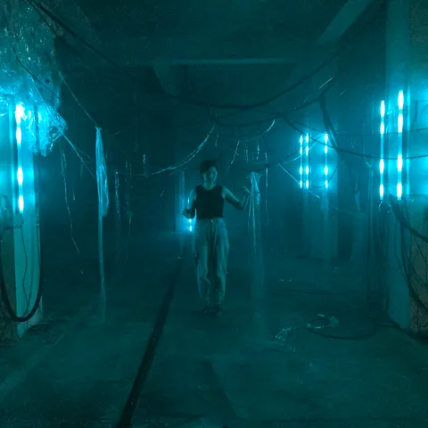 A person stands in a dimly lit room with glowing blue lights, surrounded by hanging wires and plastic sheets, creating an industrial ambiance.