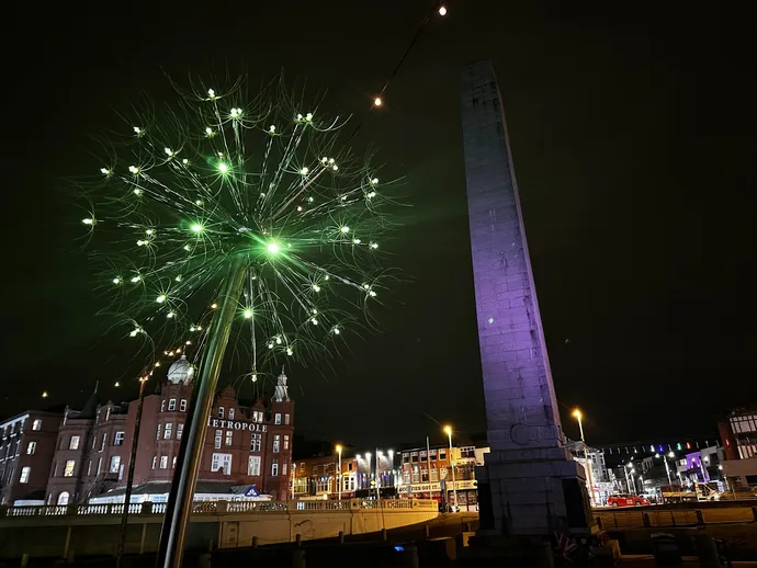 A glowing green dandelion-like light installation next to a tall stone monument in a city square at night.