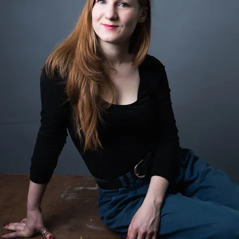 A woman with long red hair, wearing a black top and blue pants, sits on a wooden surface against a gray background, smiling slightly.