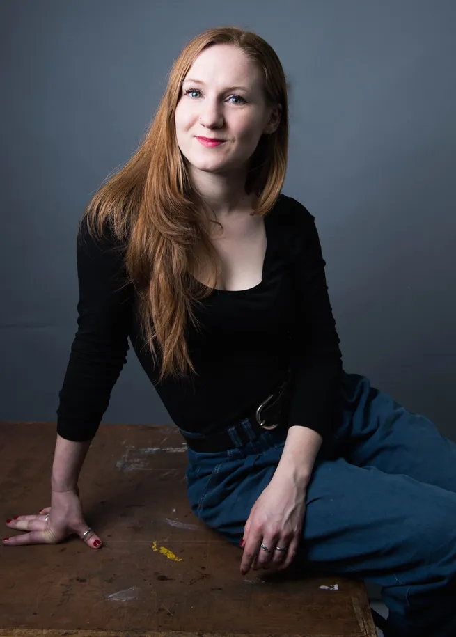 A woman with long red hair, wearing a black top and blue pants, sits on a wooden surface against a gray background, smiling slightly.