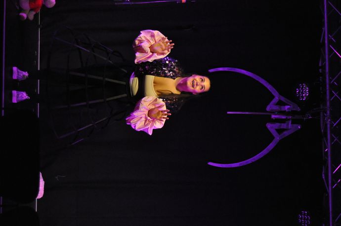 A performer on stage with a black background, wearing a unique outfit with large pink sleeves and a cage-like skirt, lit by purple lighting.