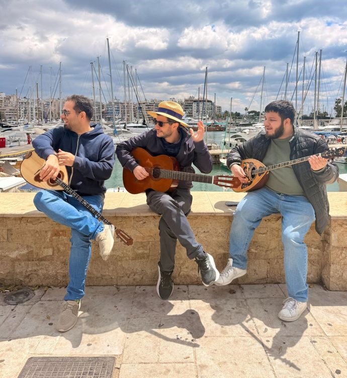 Three men play string instruments by a marina under a partly cloudy sky, creating a lively, musical atmosphere.