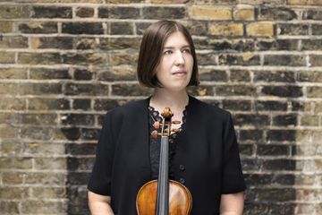 A woman with brown bobbed hair in a black outfit holds a violin in a room with a brick background