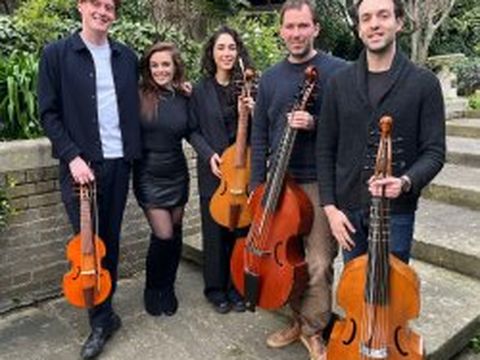 Five people standing outdoors, holding string instruments. Three men and two women, smiling, with greenery and steps in the background.