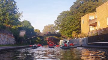 People kayaking on a canal under a bridge, surrounded by brick buildings and trees on a sunny day.