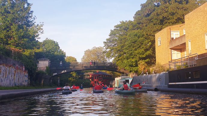 People kayaking on a canal under a bridge, surrounded by brick buildings and trees on a sunny day.