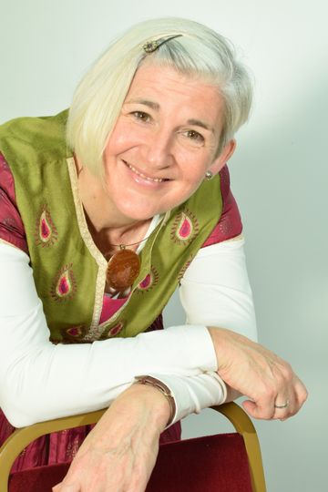 A person with short light-colored hair leans on the back of a chair, wearing a patterned vest and a pendant necklace against a plain background.