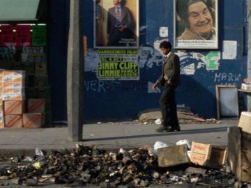 A person walks past a wall with posters and graffiti, next to a heap of trash and boxes on an urban street.