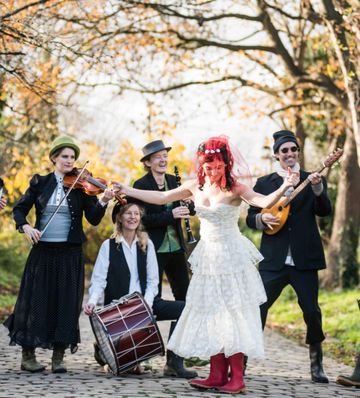 A lively band performs outdoors, featuring a violinist, drummer, and guitarist. The central figure wears a white dress and red boots, smiling brightly.