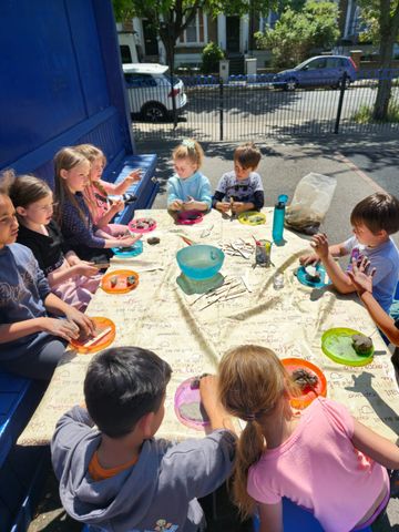 Children seated around a table outdoors, engaging in a craft activity with sticks and clay, under bright daylight.