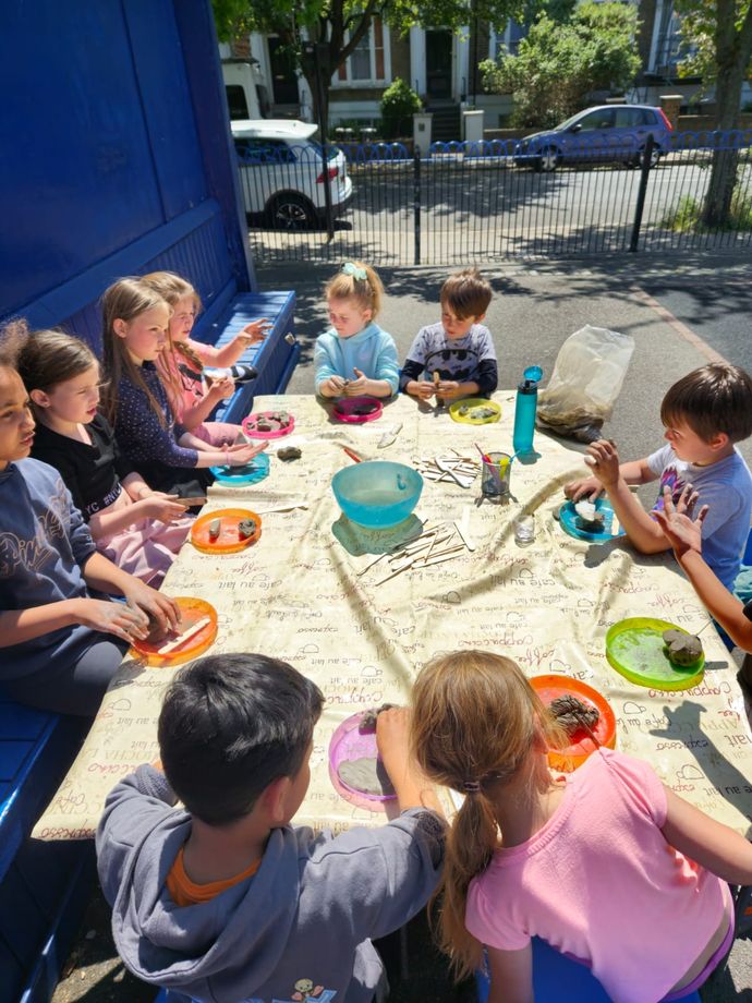 Children seated around a table outdoors, engaging in a craft activity with sticks and clay, under bright daylight.