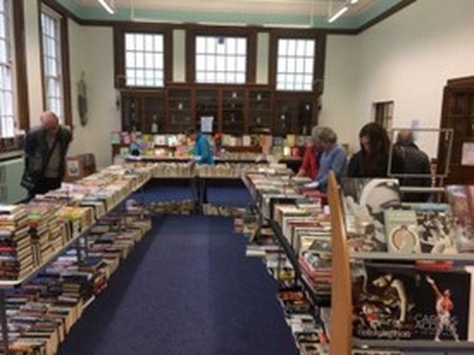 A group of people browse tables filled with books inside a bright, spacious room with tall windows and wooden cabinets.