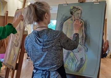 Artists in a studio drawing a live model on large easels, with focus on a woman sketching a figure on blue paper.