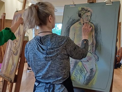 Artists in a studio drawing a live model on large easels, with focus on a woman sketching a figure on blue paper.