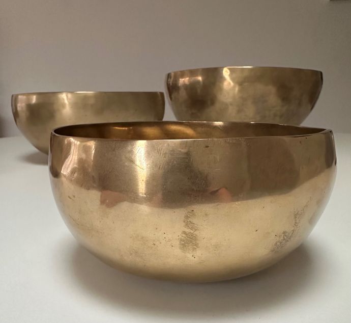 Three polished, round brass bowls of varying sizes are arranged on a smooth surface, reflecting light in a soft, warm glow.