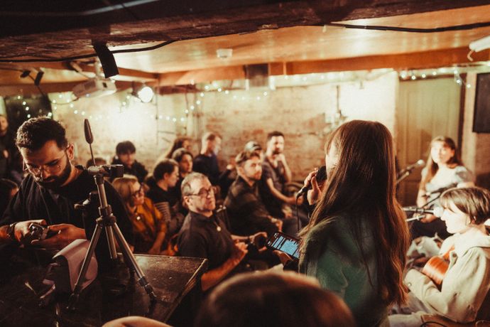 A cozy room filled with people listening to a live acoustic music performance. Strings of lights add a warm ambiance.