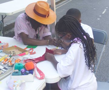 A woman and two children sit at a table outdoors, engaged in a craft activity with colorful yarn, thread, and fabric.