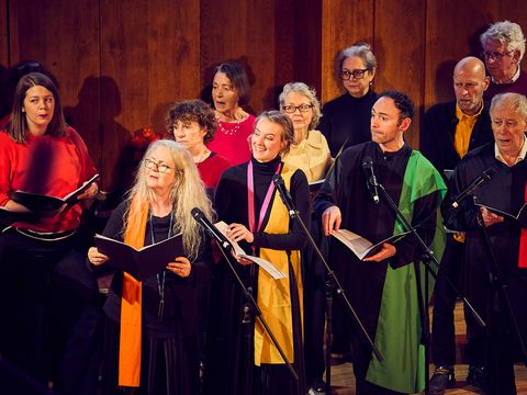 A choir of diverse individuals sings on a wooden stage, holding sheet music and wearing colorful scarves and garments.