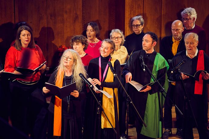 A choir of diverse individuals sings on a wooden stage, holding sheet music and wearing colorful scarves and garments.