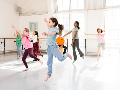 Children in casual clothes leap energetically in unison during a dance class in a brightly lit studio with large windows.