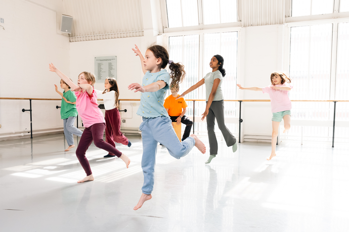 Children in casual clothes leap energetically in unison during a dance class in a brightly lit studio with large windows.