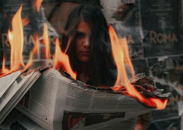 An opened newspaper burning with large flames, with a person visible in the background through the fire.