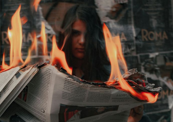 An opened newspaper burning with large flames, with a person visible in the background through the fire.