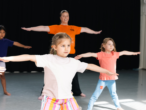 Children practicing stretching exercises with outstretched arms in a dance studio, led by an instructor wearing an orange shirt.