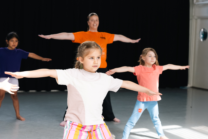 Children practicing stretching exercises with outstretched arms in a dance studio, led by an instructor wearing an orange shirt.