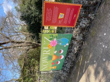 Banner on a park fence with "Nowruz" and spring illustrations, alongside a red informational sign. Trees and path in the background.