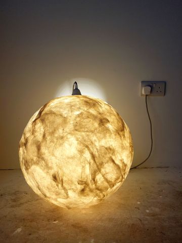 A spherical lamp with a textured, glowing surface resembling a moon, placed on a floor against a plain wall with a visible power outlet.