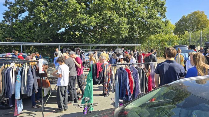 Busy outdoor clothing market with shoppers browsing racks, cars parked, and trees surrounding area.
