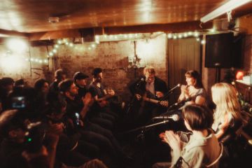 Small intimate concert in basement with audience seated closely, string lights, acoustic guitarist performing live.