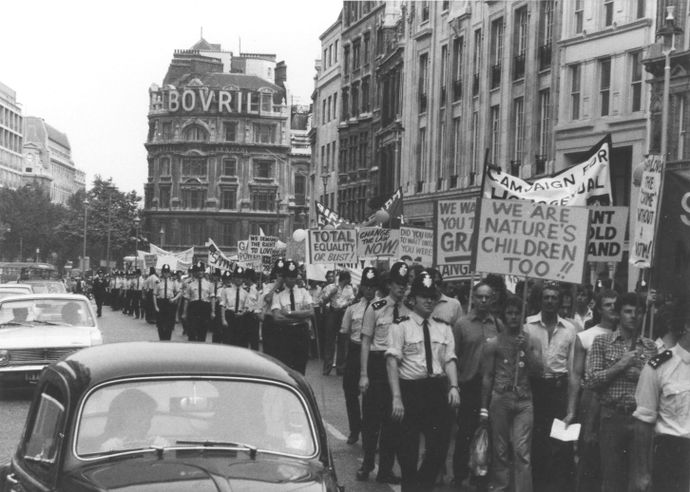 Black and white photo showing police and civilians gathering on a busy London street carrying placards handwritten with slogans like 'TOTAL EQUALITY OR BUST!'