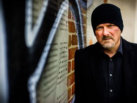 Man in a black beanie and coat leans against a graffiti-covered brick wall, looking at the camera.