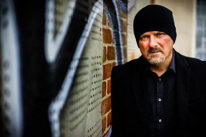 Man in a black beanie and coat leans against a graffiti-covered brick wall, looking at the camera.