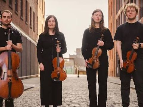 4 individuals wearing all black outfits holding 3 violin and 1 guitar standing in alleyway