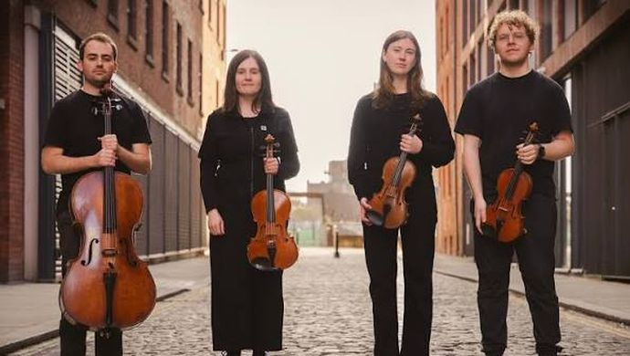 4 individuals wearing all black outfits holding 3 violin and 1 guitar standing in alleyway
