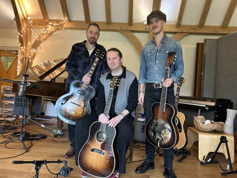 Three musicians with acoustic guitars posing in studio, piano background, microphones, warm wooden interior setting.