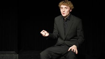 Person in black suit seated on stage, gesturing while performing under dramatic spotlight lighting.