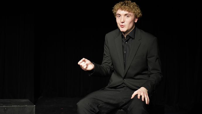 Person in black suit seated on stage, gesturing while performing under dramatic spotlight lighting.