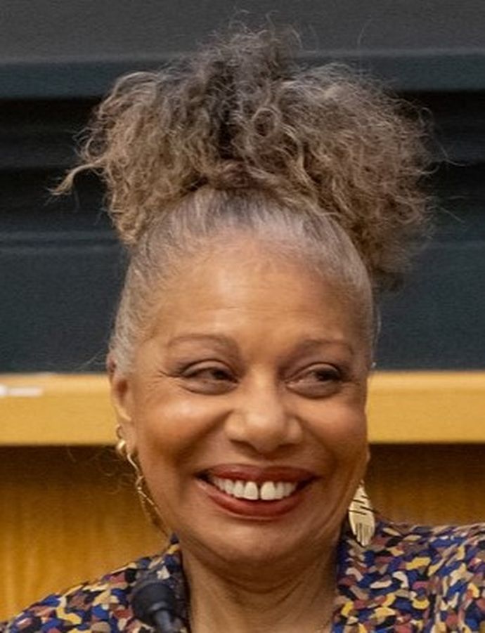 Smiling person with curly hair in a bun, wearing a patterned top and gold earrings, sitting in front of a dark background.