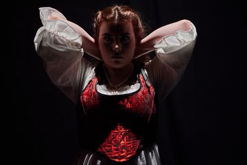 Woman with brown hair, middle parted, stands with hands behind head, wearing white gown with red bodice over it. Serious expression with face in shadow