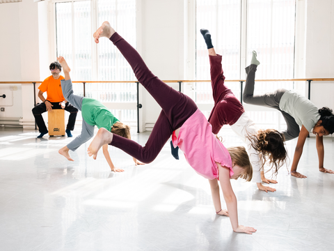 Children in colorful clothes performing handstands in a bright dance studio, with a person playing a cajón drum in the background.
