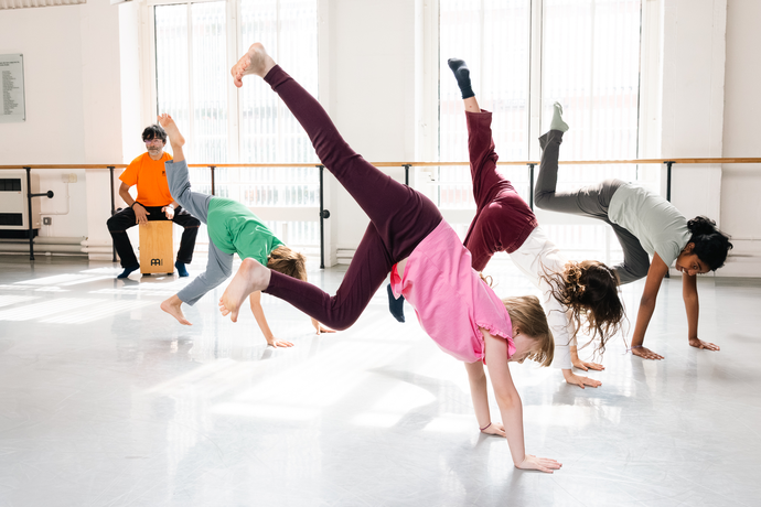 Children in colorful clothes performing handstands in a bright dance studio, with a person playing a cajón drum in the background.