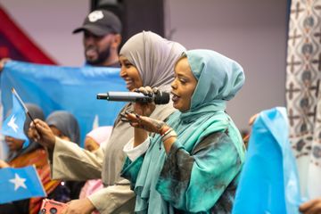 Two women in colorful hijabs sing into microphones, surrounded by people holding blue flags with white stars.