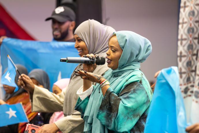 Two women in colorful hijabs sing into microphones, surrounded by people holding blue flags with white stars.