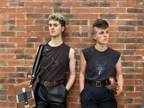 Two individuals with short hair lean against a brick wall, both wearing sleeveless shirts. One holds a violin and the other a wooden instrument case.